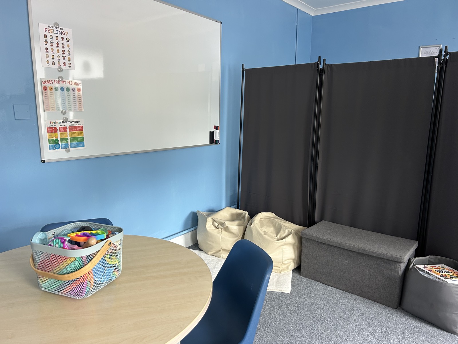 A round table and whiteboard with feelings charts, alongside bean bags and a basket of sensory toys